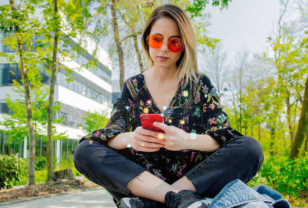Fiatal nő napszemüvegben mobiltelefonnal ül a zöld sikátorban, közel az egyetem modern épületéhez  - Fotó, kép