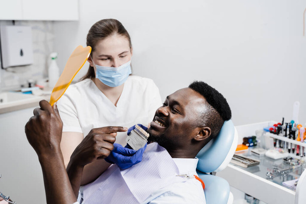 歯だ。歯のクリニックでアフリカ人患者の歯のホワイトニングのための歯の色の色合いガイドを示す歯科医。アフリカ人は、医師の手でベンダーやインプラントの歯の色マッチングサンプルを見ています - 写真・画像