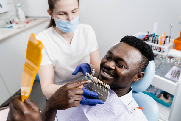 Dentistry. Doctor checking teeth color matching samples in dental clinic. Teeth whitening of african man. Dentist working with teeth color shades guide - Fotografie, Obrázek