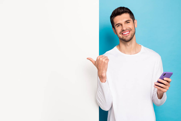 Photo of handsome positive man hold telephone direct finger empty space isolated on blue color background. - Fotó, kép