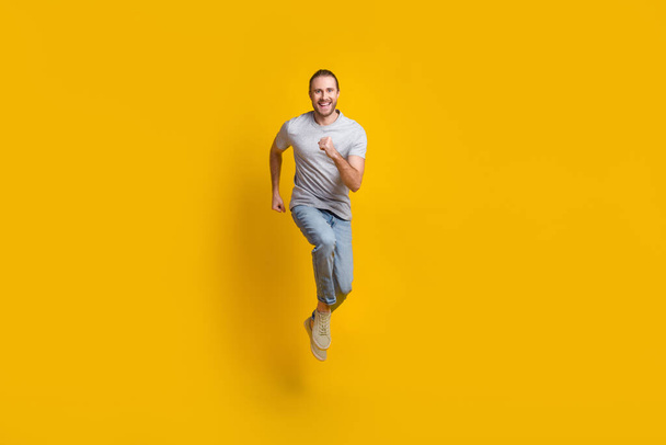 Full length photo of pretty impressed man wear grey t-shirt running fast jumping high isolated yellow color background. - Foto, Imagen