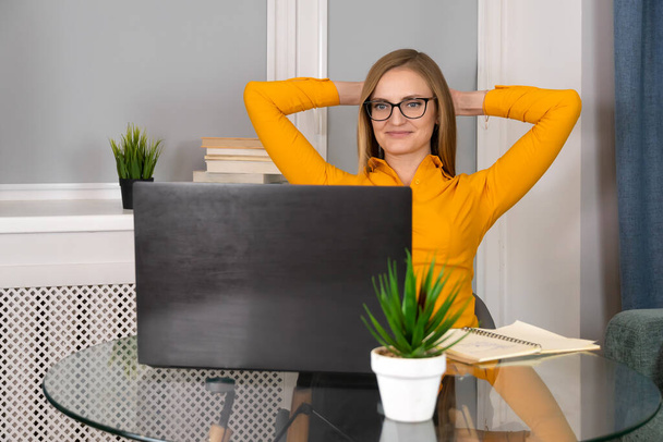 Close-up of a smiling woman in an orange blouse and glasses sitting at a glass table, puts her hands behind her head and looks at a laptop. Concept of success, joy, victory, rest - Fotoğraf, Görsel