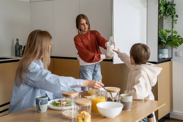 Napi családi reggeli rutin. Anya, lánya és fia együtt reggeliznek, a konyhaasztalnál ülnek, gabonapelyhet esznek tejjel és narancslevet isznak. Családi étkezési idő fogalma - Fotó, kép