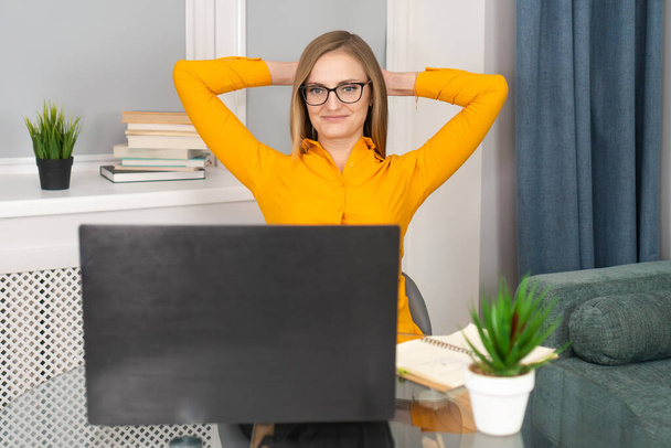 Happy woman in an orange blouse and glasses sits at a glass table, puts her hands behind her head and looks at her laptop with a smile. Concept of success, joy, victory, rest - Foto, afbeelding