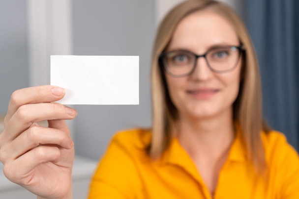 Close-up of a white business card in the hand of a woman with glasses and an orange shirt. Specialist offering his services - Фото, изображение