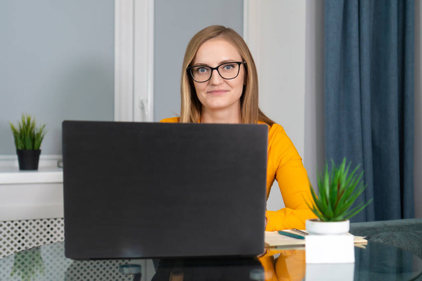Smiling woman with glasses sits at a table with a laptop in a cozy room and looks into the camera, next to a business card with a place for text. Professional, specialist - Foto, imagen
