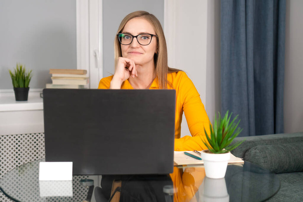Successful smiling business woman in glasses and an orange shirt using a laptop looks at the camera. Business, psychology, work, advertising - Photo, image
