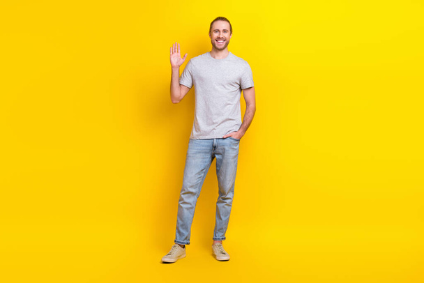 Full size photo of good mood friendly man wear gray t-shirt jeans hand in pocket waving palm say hi isolated on yellow color background. - Foto, afbeelding