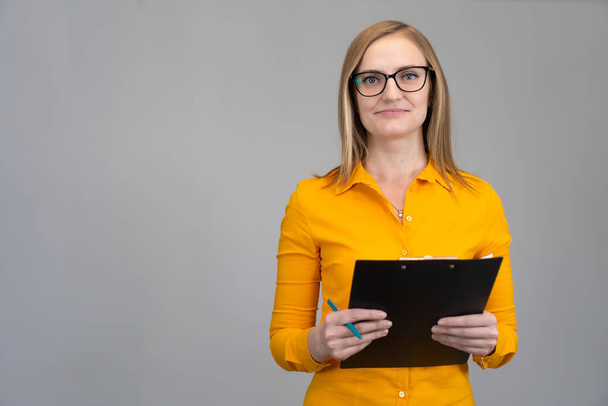 Smiling business woman in an orange shirt and glasses with a paper tablet in her hands against a light wall, copying the space. Business concept, achievements, career, wealth, training, advertising - Фото, изображение