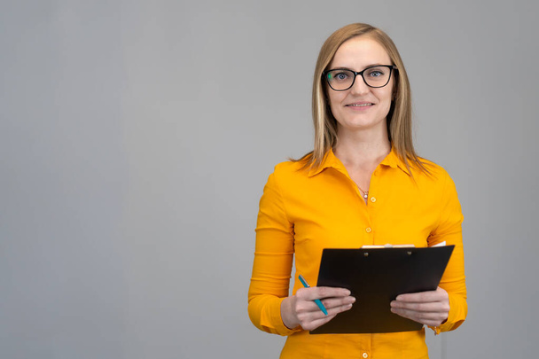 Smiling business woman in an orange shirt and glasses with a paper tablet in her hands against a light wall, copying the space. Business concept, achievements, career, wealth, training, advertising - Φωτογραφία, εικόνα