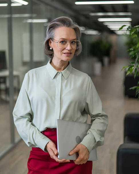 Mature caucasian woman stands with a laptop among the office - Фото, изображение