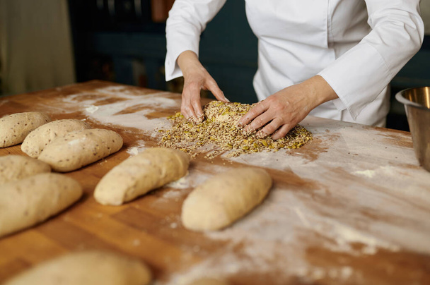 Retrato de panadero femenino positivo que forma panecillos, sumergiendo la masa con semillas y copos de avena. Panadería concepto de casa - Foto, imagen