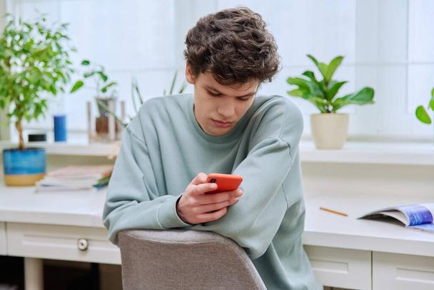 Jóképű fiatal srác okostelefonnal ül a széken, közel az ablakhoz otthon. Férfi SMS-t ír a telefonján. Mobil alkalmazások, internetes online technológiák szabadidős tanuláshoz csevegés - Fotó, kép