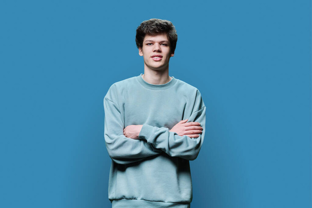 Portrait of young confident male 18-20 years old in casual sweatshirt, with crossed arms on blue studio background. Handsome guy with curly hair looking at camera. Lifestyle, youth concept - Photo, Image