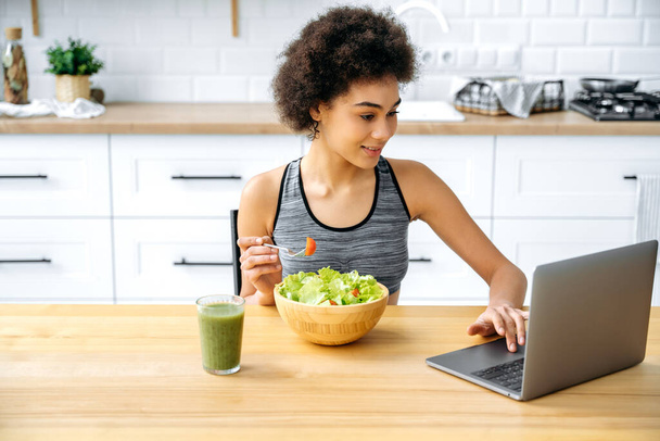 Pozitivní Afroameričanka sedí doma u stolu v kuchyni, jí čerstvý zeleninový salát, dívá se na video na notebooku o zdravém stravování, stravě, on-line cvičení videa, úsměvy - Fotografie, Obrázek