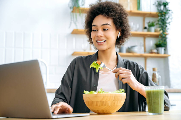Gezond levensstijl concept. Moderne positieve Afrikaanse Amerikaanse jonge vrouw, zitten aan tafel in de keuken, het eten van verse groente salade, kijken naar gezond eten of thuis workout video 's op laptop, glimlachen - Foto, afbeelding
