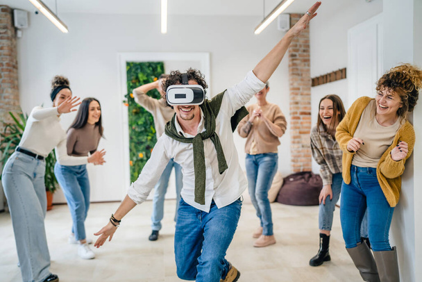 One man adult caucasian male in front of group of men and women friends enjoy virtual reality VR headset at work having fun together during team building seminar real people bright filter - Foto, imagen