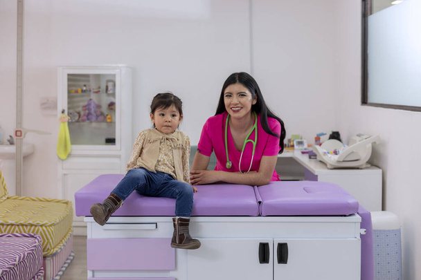 Portrait of a pediatrician and a little girl on the table in their medical office looking at the camera, with copy space - Fotó, kép
