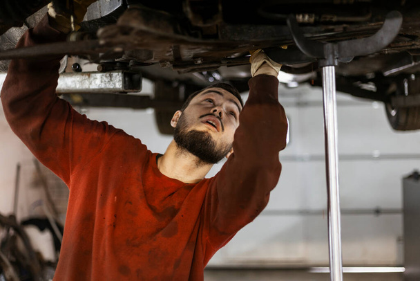 Un mecánico trabajador con ropa manchada de aceite está trabajando debajo de la parte inferior de un coche, desmontando y quitando un bastidor de dirección. - Foto, imagen