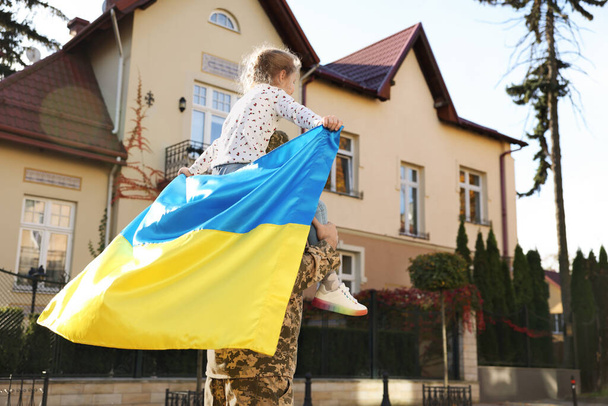 Soldier in military uniform carrying his daughter on shoulders and Ukrainian flag on city street, space for text. Family reunion - Fotoğraf, Görsel