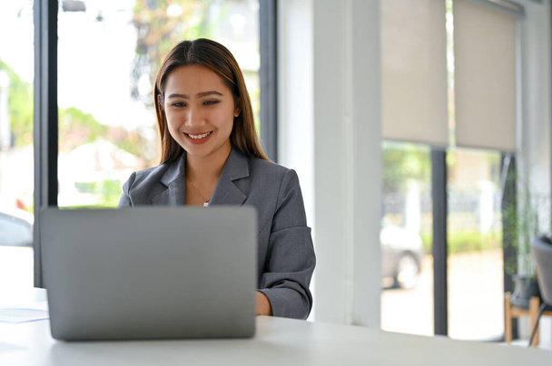 Attractive and smiling millennial Asian businesswoman in formal business suit using her laptop at a table in the office. - Photo, Image