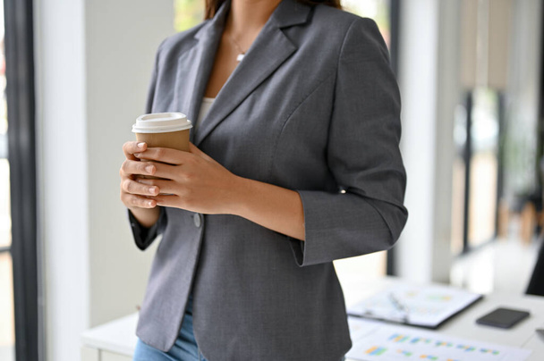 Image recadrée d'une femme asiatique magnifique et confiante PDG ou patron en costume d'affaires formelle se tient dans son bureau et tient une tasse de café à emporter. - Photo, image