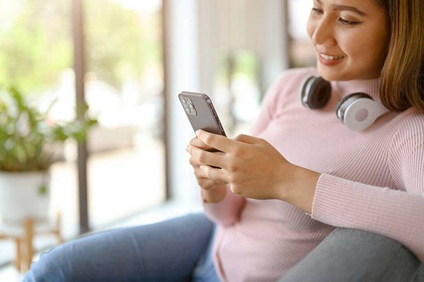 Close-up image of a beautiful and happy millennial Asian woman in causal clothes using her smartphone while relaxing in living room. - Foto, imagen