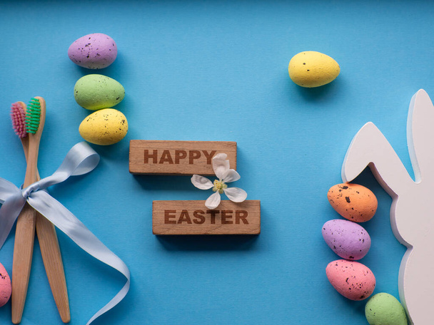 Happy Easter and dentist concept. Wooden toothbrushes with Easter decorations on blue background. Dentist Easter greating card. Top view, flat lay. Bamboo toothbrushes, colorful eggs, white bunny. - Photo, image