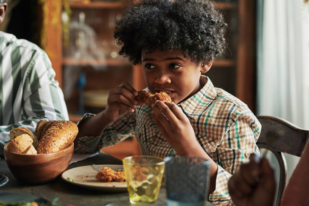 African little boy sitting at dining table and eating roast chicken during dinner with his family - Photo, Image