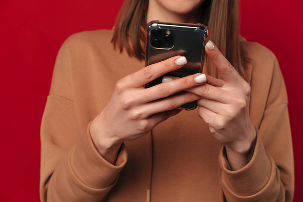 Studio plan sans visage d'une jeune femme tenant et tapant à son téléphone sur fond rouge. - Photo, image