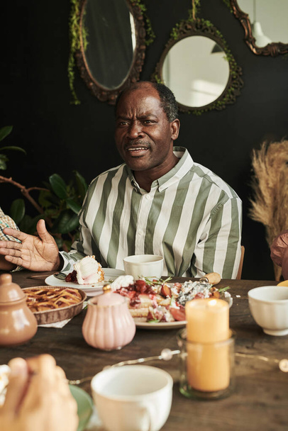 Afričan dospělý muž sedí u stolu a mluví při jídle dort a pití čaje během sváteční příležitosti - Fotografie, Obrázek