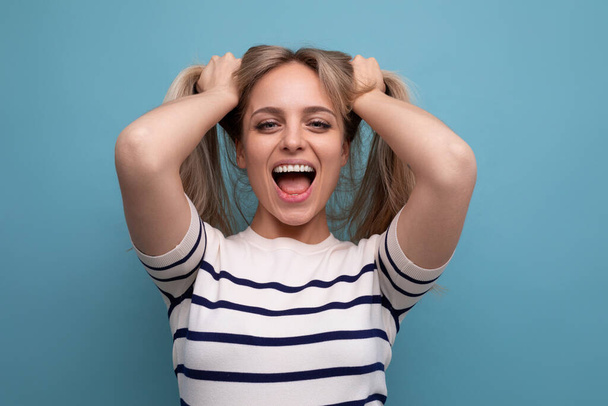 cheerful blond young woman holds her hair in her hands and laughs cutely on a blue background. - Photo, Image