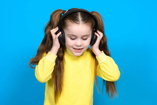 A little girl in a yellow sweater on a blue background listens to music in wireless headphones and holds them in her hands. - Foto, afbeelding