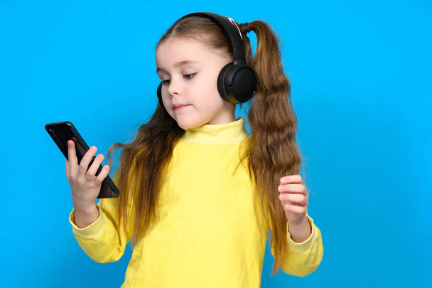 Portrait of a little girl with a phone and wireless headphones, a child in a yellow sweater on a blue background, a child dances when he hears music. - Photo, Image