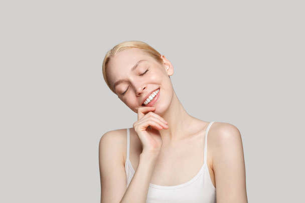 Glückliche schöne blonde Frau berührt ihr Gesicht und lächelt isoliert auf grauem Hintergrund. Schönheit, Kosmetik, Hautpflege, Glamour - Foto, Bild