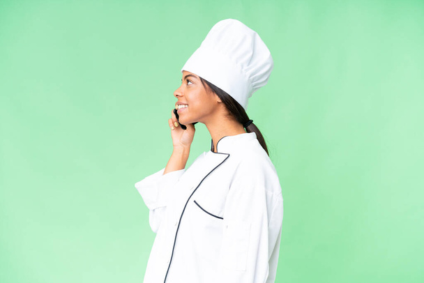 Jonge Afro-Amerikaanse chef-kok over geïsoleerde chroma key achtergrond houden van een gesprek met de mobiele telefoon met iemand - Foto, afbeelding