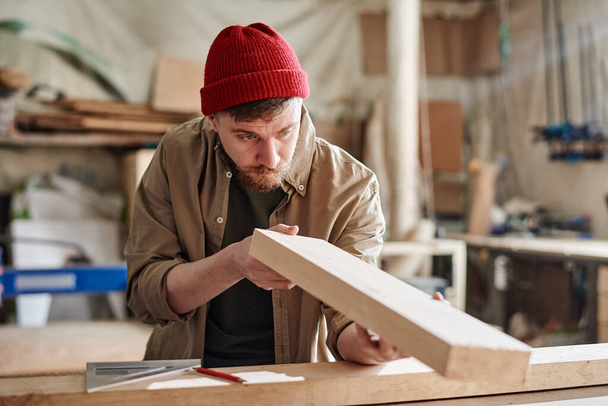 Retrato horizontal medio del joven hombre caucásico que trabaja en taller de carpintería revisando la superficie de madera del tablón antes de pulirlo - Foto, imagen