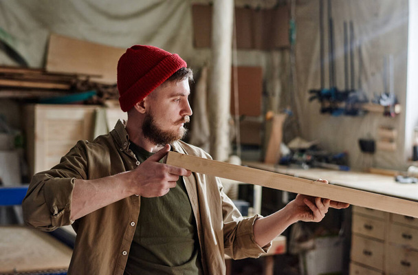 Young male Caucasian carpenter with beard on face wearing red knit cap looking at wooden plank surface to check its quality - Photo, Image
