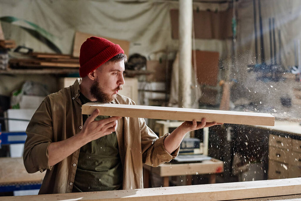 Современный плотник в красной вязаной кепке держит деревянную доску, выдувая пыль из нее в столярной мастерской. - Фото, изображение