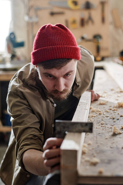 Verticaal schot van serieuze jonge mannelijke timmerman met behulp van stalen vierkant bij het werken met houten plank in houtwerkplaats - Foto, afbeelding
