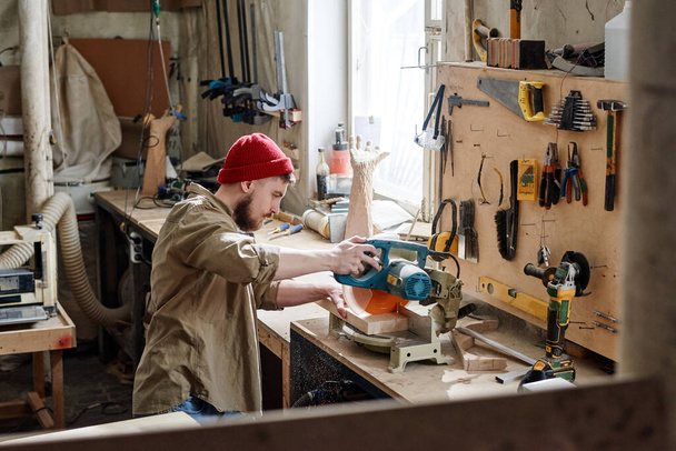 Alto ângulo de jovem carpinteiro caucasiano corte pedaço de prancha de madeira com serra de mitra na oficina - Foto, Imagem