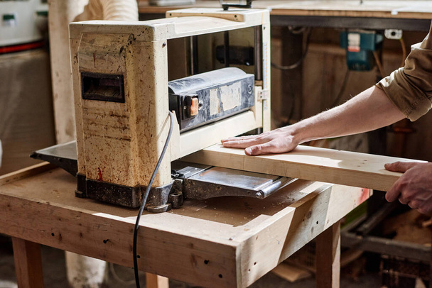 Μη αναγνωρίσιμος αρσενικός ξυλουργός που εργάζεται χρησιμοποιώντας μηχανή πλανητών πάχους για να κόψει τη σανίδα ξύλου στο εργαστήριο ξυλουργικής - Φωτογραφία, εικόνα