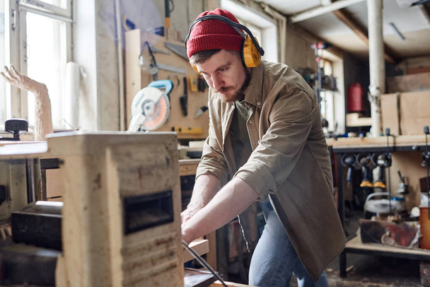Young male carpenter wearing noise-cancelling headphones using thickness planer machine in woodworking workshop - Foto, Bild