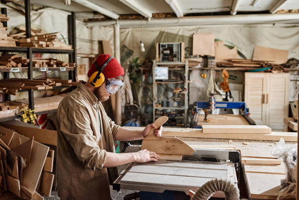 Jovem carpinteiro masculino vestindo óculos de segurança e fones de ouvido de proteção auditiva cortando prancha de madeira com serra de mesa - Foto, Imagem
