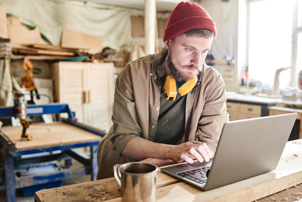 Boynunda kulaklık olan sakallı genç marangoz kahve molası sırasında dizüstü bilgisayarda internette geziniyor. - Fotoğraf, Görsel