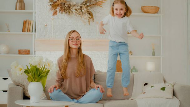 Mindful calm Caucasian mother in glasses meditating sitting on sofa while active noisy child daughter jumping on couch. Peaceful mom relaxing in yoga meditation at home naughty little kid girl jump - Fotografie, Obrázek
