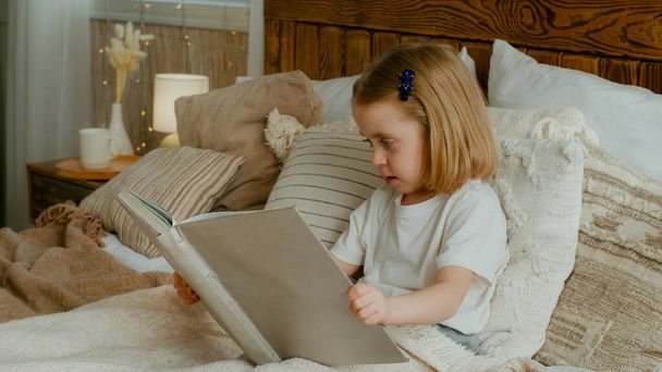 Cute little 5s Caucasian alone child girl daughter looking in book kid preschool baby sitting in bed watching pictures in copybook studying learning read try reading elementary education in bedroom - Fotografie, Obrázek