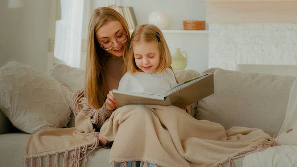 Caring single parent Caucasian mother babysitter mom hug little preschool child girl kid daughter covered with blanket sit on couch learn read book fairy tale story bonding family education at home - Fotó, kép