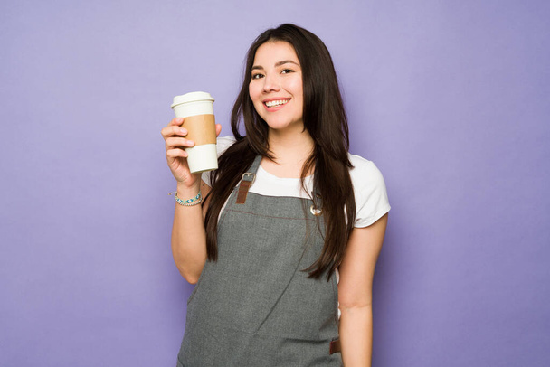 Χαρούμενη νεαρή γυναίκα με ποδιά που εργάζεται ως μπαρίστα χαμογελώντας ενώ σερβίρει καφέ για να πάει στο καφέ - Φωτογραφία, εικόνα