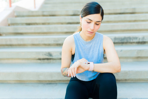 Mujer atractiva en forma hispana usando la aplicación fitness en su smartwatch después de hacer ejercicio y correr al aire libre - Foto, Imagen